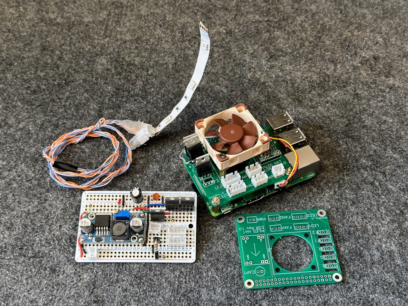 Prototype circuit board, finished Raspberry Pi and green custom HAT with a small Noctua fan, LED strip and wiring, plus a spare bare PCB laid on grey carpet.